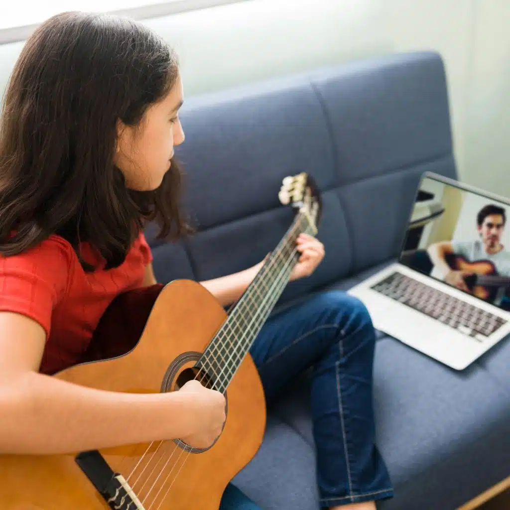 hispanic girl with music skills listening instructions her guitar teacher video call laptop while sitting sofa home 1 1024x1024